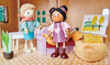 Two Ayana and her Cat figures in a cozy room, one seated on a sofa wearing a pink dress and headband, the other standing in blue shorts and a green jacket, surrounded by quaint home decor and a plush