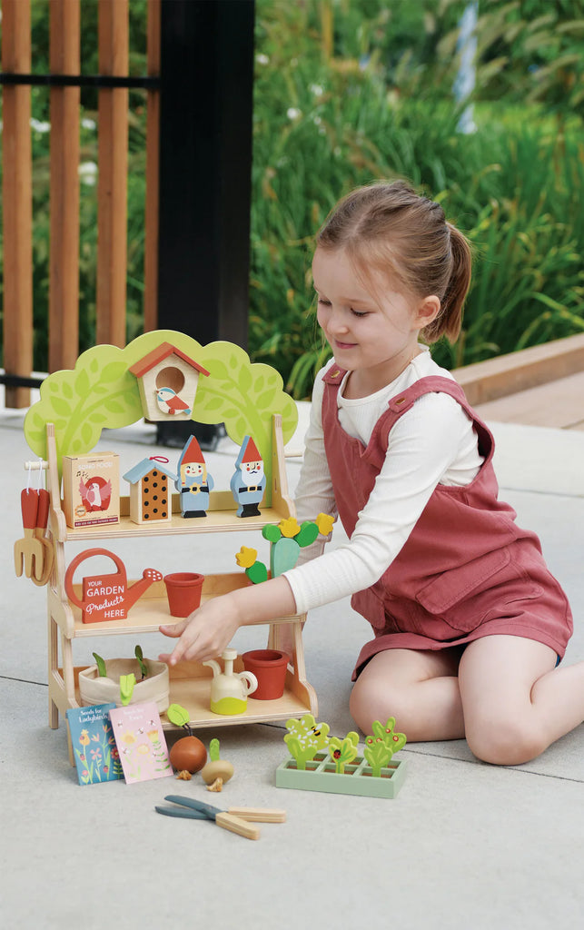 A young child in a pink romper and white shirt enjoys pretend play with the Garden Center, featuring potted plants, tools, a bee house, and figurines. This outdoor activity on a sunny day enhances social and language skills in a vibrant garden setting.