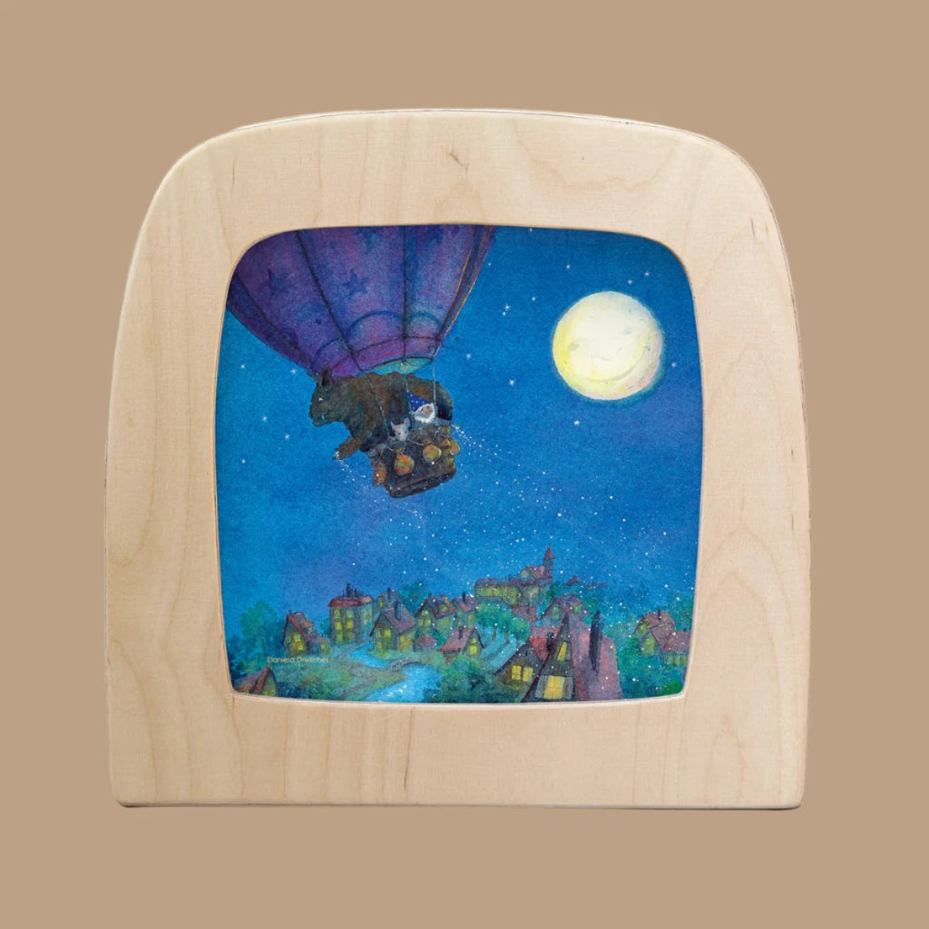 A whimsical scene featuring the "Goodnight Sandman" silhouette, showcases bears in a hot air balloon floating over a village at night, framed by a magic window with a large moon illuminating the starry sky for the Toverlux Lamp.