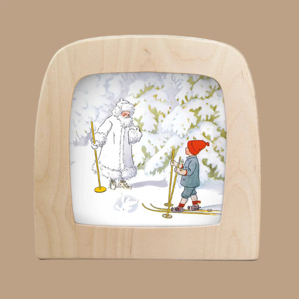 The framed "Silhouette Only - Ollie's Ski Trip silhouette - Jack Frost for Toverlux Lamp" depicts a child in a red hat and coat with ski poles, facing a snow creature on skis amidst magical silhouettes, snow, and trees. The frame is crafted from light wood.