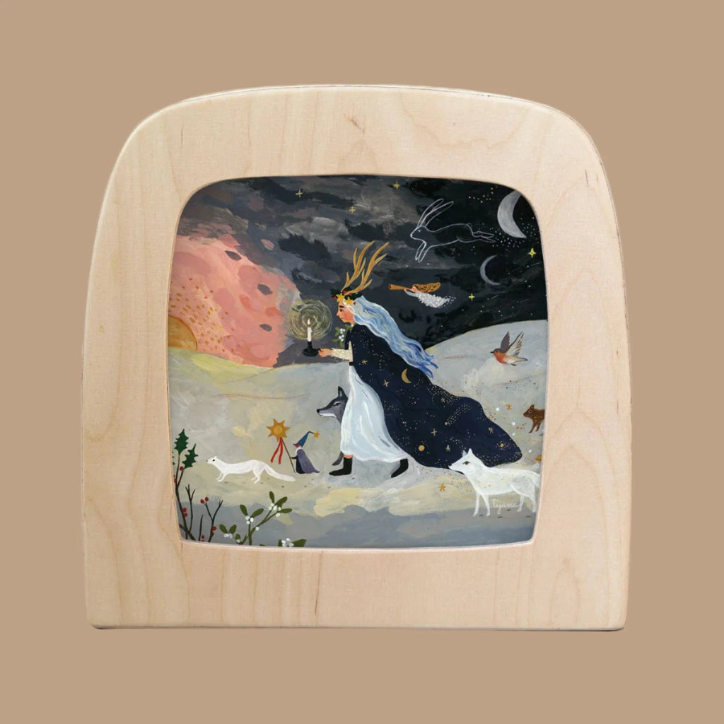 A wooden frame displays "Silhouette Only - Winter Solstice For Toverlux Lamp," depicting a figure with flowing hair and a starry cloak, holding a lantern amid white animals on a dreamy landscape under birds, the moon, and a pink sun. Ships in one week.