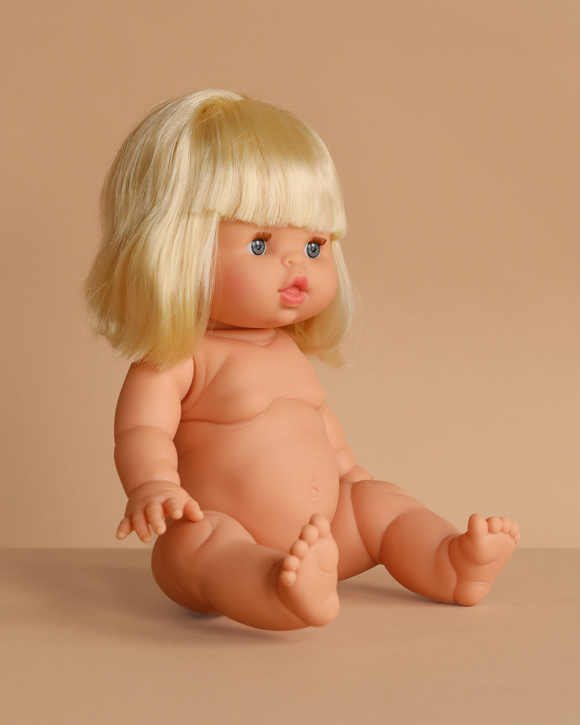 The Minikane Baby Doll (13") - Angela, anatomically correct with blonde hair and blue eyes, sits nude on a beige surface against a beige background, facing slightly right. Clothing sold separately. Made in Spain.