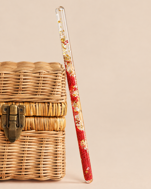 A clear, cylindrical Konges Sloejd Magical Glitter Wand - Cherry filled with red beads, gold glitter, and small cherry-shaped charms, leaning against a woven basket.