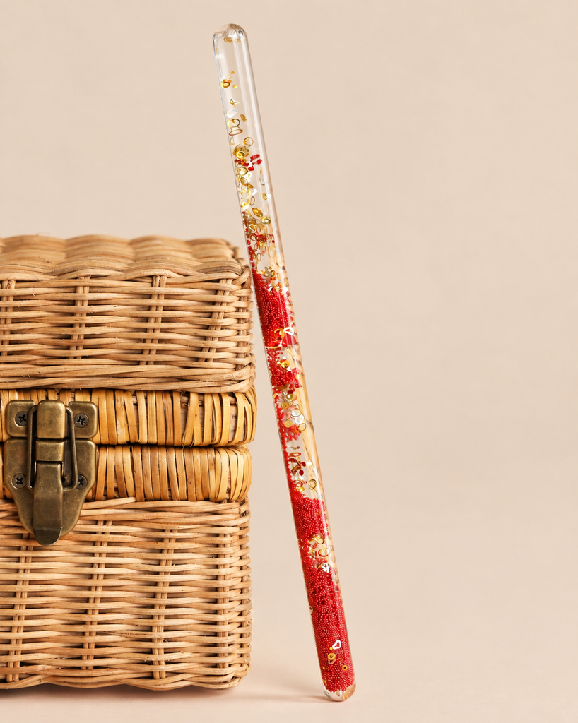 A clear, cylindrical Konges Sloejd Magical Glitter Wand - Cherry filled with red beads, gold glitter, and small cherry-shaped charms, leaning against a woven basket.