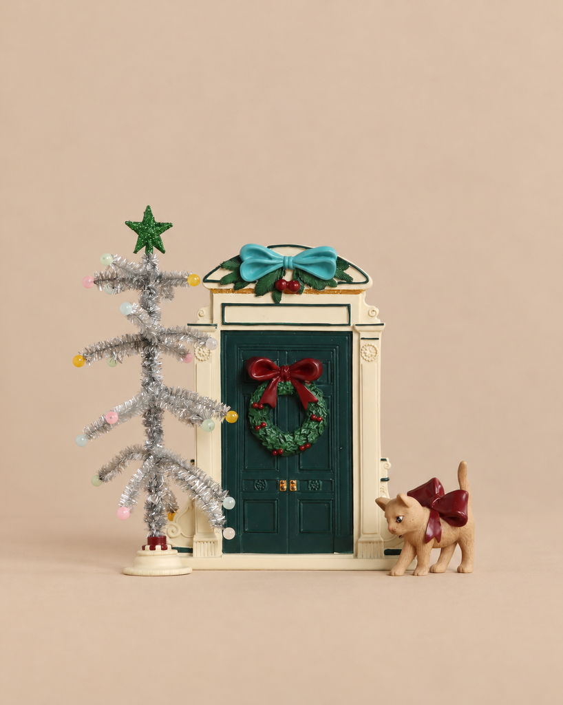 A Konges Sloejd Christmas Door featuring a miniature green elf door topped with a blue bow, a silver tinsel tree with colored lights, and a small cat wearing a red bow.