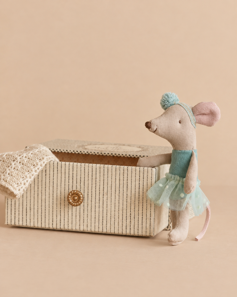 A Maileg Dance Mouse in Daybed, Little Sister - Mint stands beside a striped rectangular box with a knit blanket draped inside.