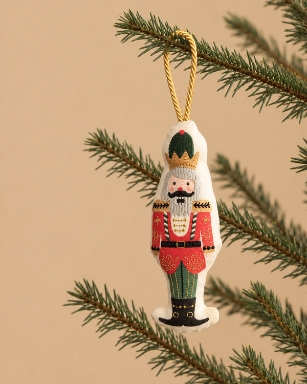 Nutcracker Embroidered Ornament hanging from a pine branch by a gold cord.