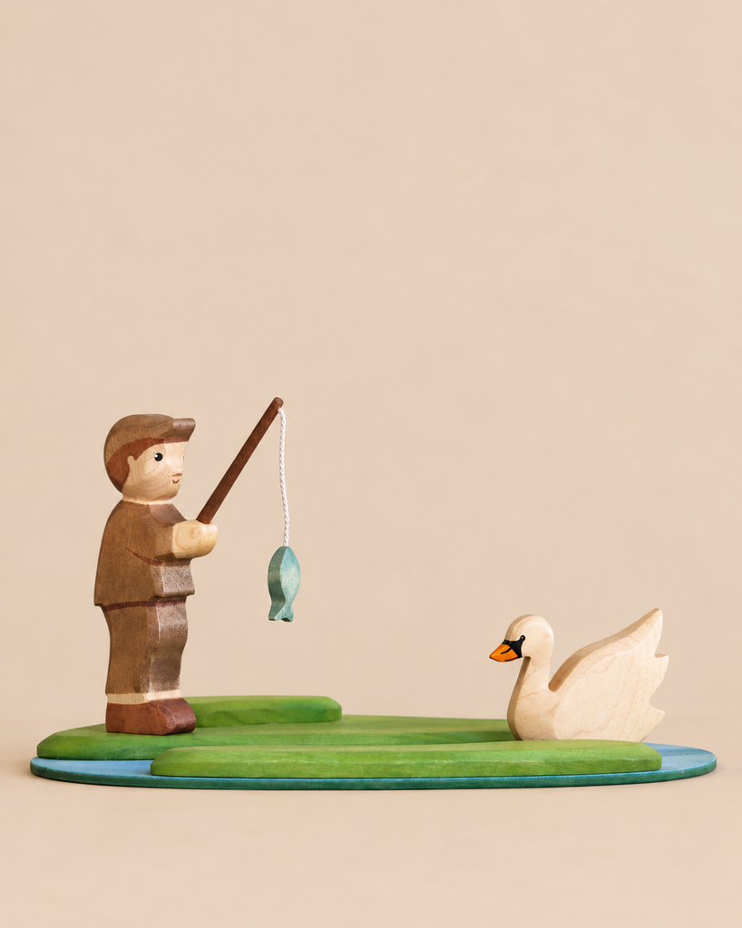 A handmade Holzwald Fishing Pond Set features a wooden toy fisherman on a green bank, casting his line toward a white swan floating on a blue pond.
