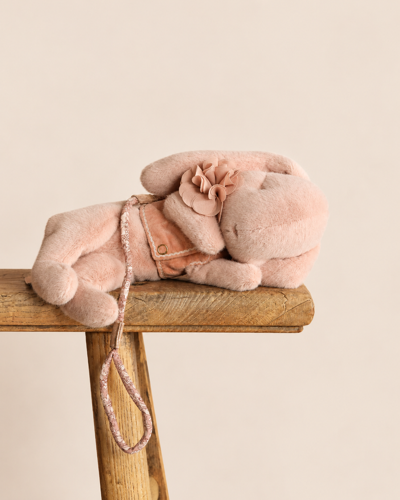 A Maileg Plush Sleeping Bunny With Harness wears a small fabric flower while lying down on a wooden stool.