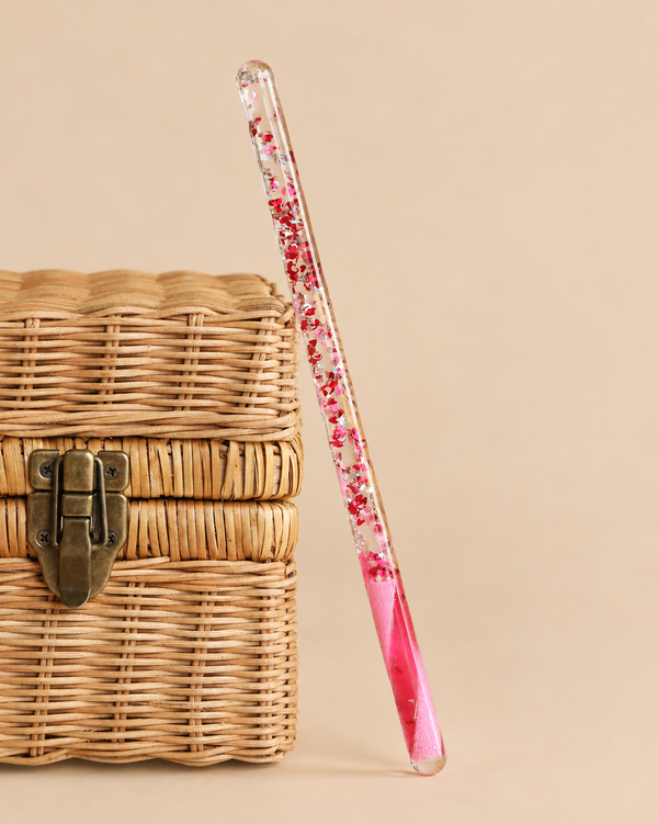 A transparent Konges Sloejd Magical Glitter Wand - Pink Mix filled with pink liquid resting against a small wicker chest.