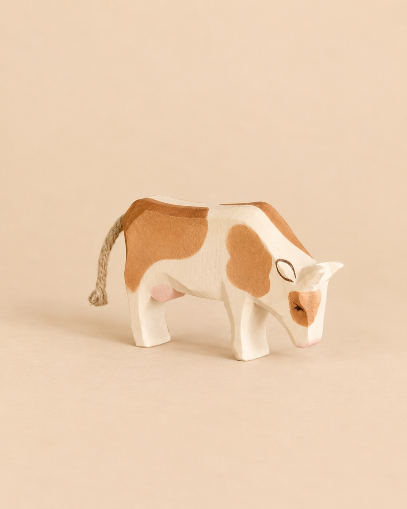 Handcrafted Ostheimer Cow - Eating, a wooden toy with brown patches, head bowed as if grazing, and a braided rope tail.
