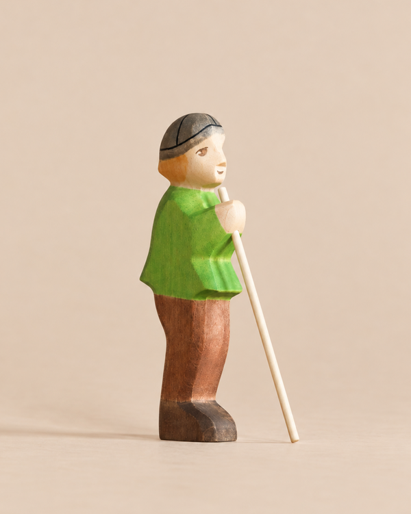 Handmade Holzwald Shepherd figurine of a person wearing a green tunic, brown trousers, and a grey cap, holding a tall staff.