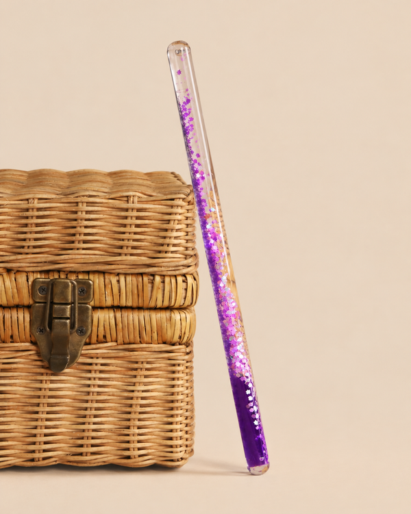A transparent Konges Sloejd Magical Glitter Wand - Unicorn filled with purple sparkles and unicorn-shaped confetti, leaning against a small woven basket.