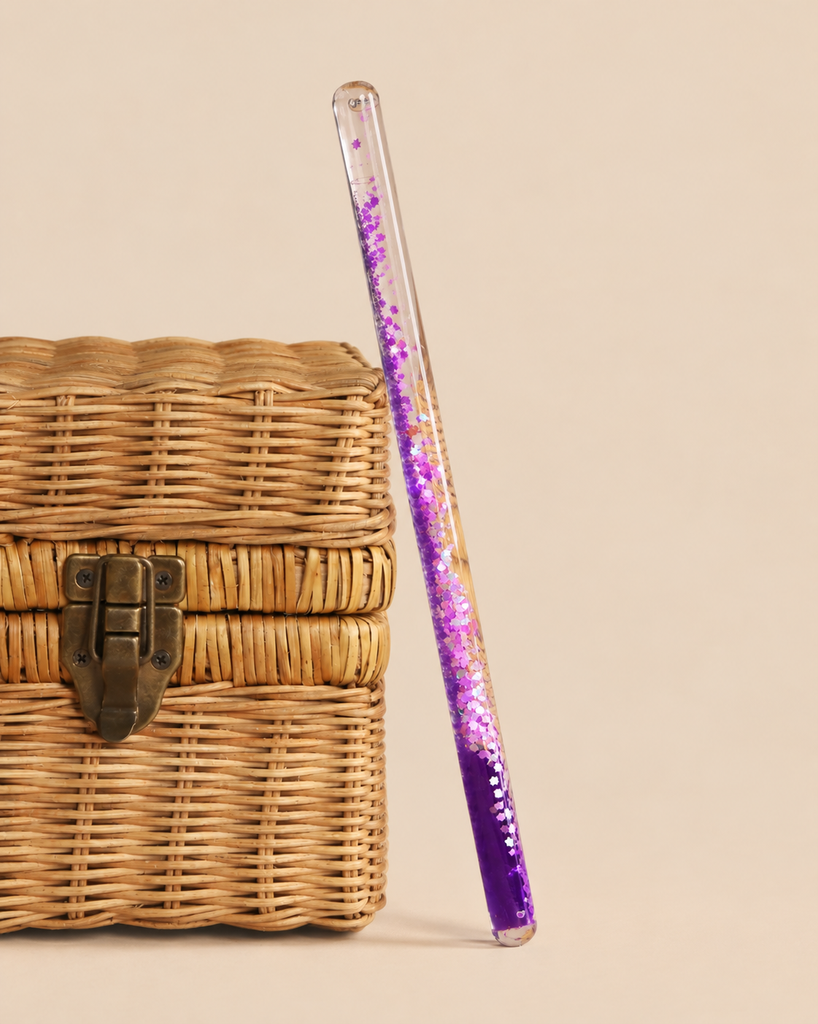 A transparent Konges Sloejd Magical Glitter Wand - Unicorn filled with purple sparkles and unicorn-shaped confetti, leaning against a small woven basket.
