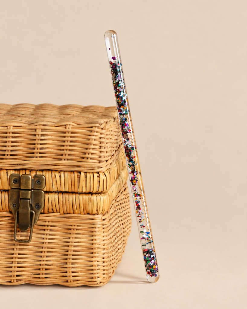 A clear Konges Sloejd Magical Glitter Wand - Star Glitter filled with multicolored star-shaped accents resting against a small wicker box.