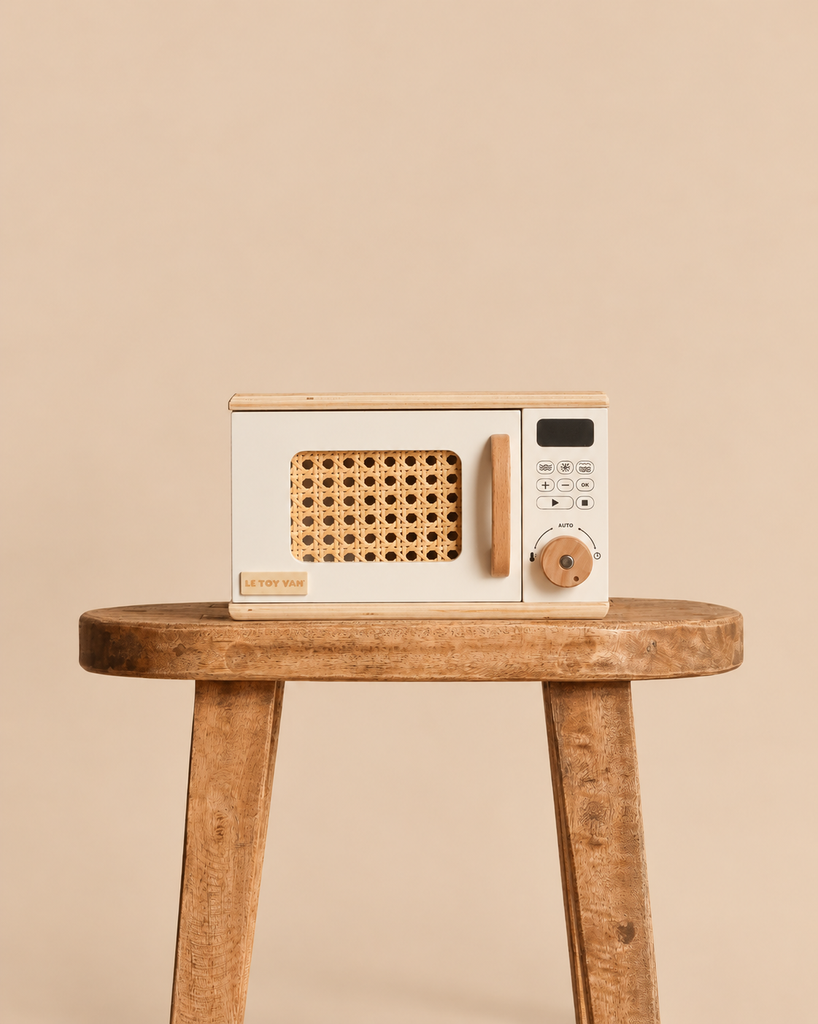 A wooden Pretend Play Microwave with a rattan door panel and wooden handle, sitting on a wooden stool as part of a play kitchen.