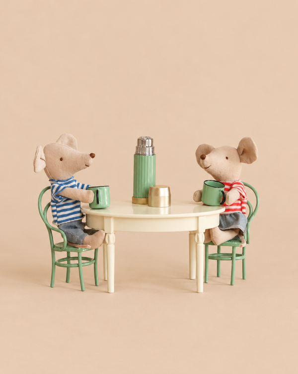 Two Maileg Little Brother and Sister Dining Set mice in striped shirts sit at a vintage dining table holding tiny green mugs, with a miniature thermos and cup between them.