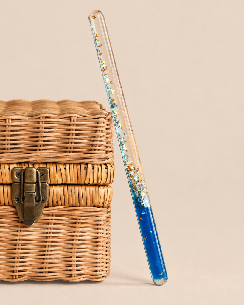 A clear Konges Sloejd Magical Glitter Wand - Rocket filled with blue liquid, stars, and moon-shaped confetti, leaning against a small woven wicker basket.