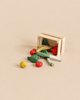A small Maileg Fruits & Veggies crate tipped on its side with miniature fruits and vegetables, including a banana, broccoli, leeks, and tomatoes, spilling out.