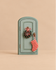 A small, arched, sage-green wooden door from the Maileg Christmas Set, decorated with a miniature wreath and a red and white striped stocking hanging from the handle.
