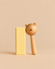 A natural Sabo Concept Bear Rattle - Natural leaning against a tall, pale yellow rectangular block.