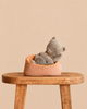 A grey Maileg Small Sleeping Kitten in Basket rests in a small, striped-lined basket perched on a wooden stool.