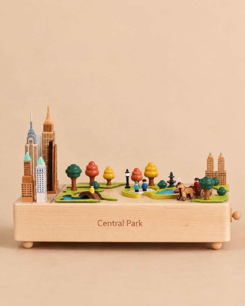 Central Park Wooden Music Box featuring a miniature scene with city buildings, trees, a pond, and tiny figures on a base labeled “Central Park.”