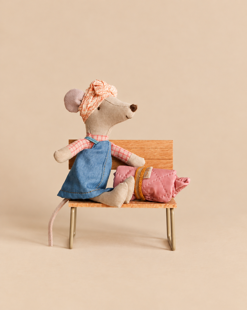 A Maileg Hiker Mouse With Blush Sleeping Bag - Big Sister sits on a miniature wooden bench, wearing a headscarf and denim overalls next to a small, rolled-up sleeping bag.