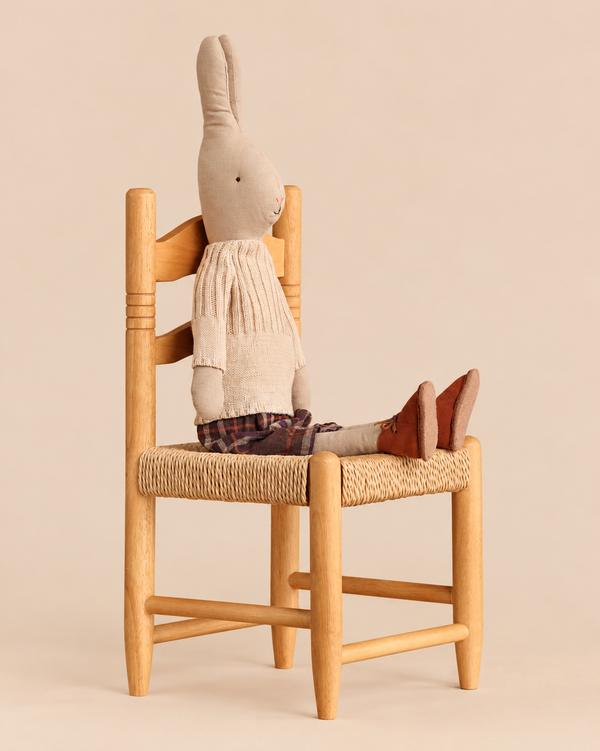 A Maileg Rabbit, Size 4 - Classic - Pants and Knitted Sweater sits upright on a small wooden chair, wearing a cream knit sweater and plaid pants.