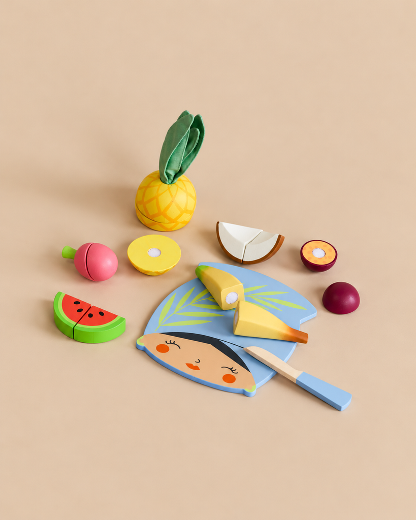 A Tropical Fruit Chopping Board shaped like a person’s face accompanies a toy fruit set including a pineapple, sliced pieces, and a toy knife for a kids play kitchen.