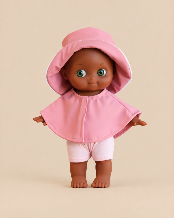 A Minikane Mini Pancha Doll - Prunelle with Pink Rain Suit, featuring a dark-skinned vinyl doll with green eyes wearing a pink sun hat, matching cape, and shorts.