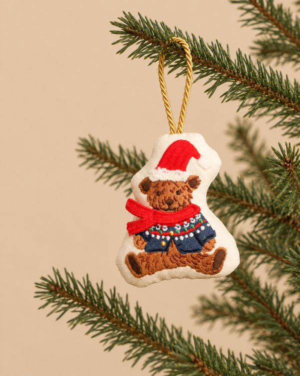 Beary Christmas Embroidered Ornament shaped like a plush cotton teddy bear wearing a Santa hat, red scarf, and blue sweater, hanging from a pine branch.