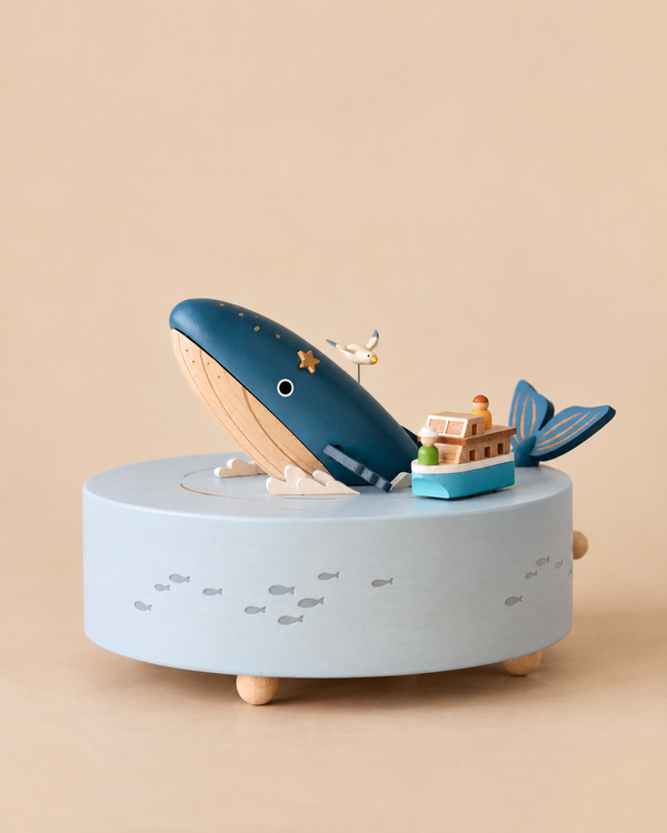 Wooden Whale Music Box featuring a blue whale, a small boat with two figures, and a seagull on a circular base decorated with fish.