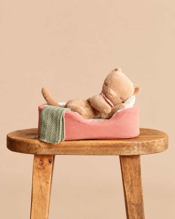 A Maileg Small Sleeping Kitten in Cozy Bed rests in its pink pet bed with a green blanket on a wooden stool.