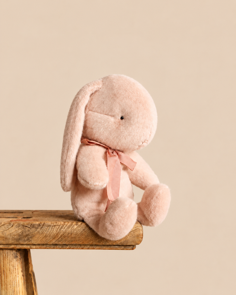 A light powder Maileg Plush Bunny, Small sits on the edge of a wooden stool, wearing a matching pink ribbon around its neck.