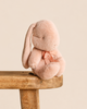 A small, pale pink Maileg Plush Bunny, Small - Powder with one long drooping ear and a tied ribbon at its neck, sitting on a wooden stool as a soft toy.