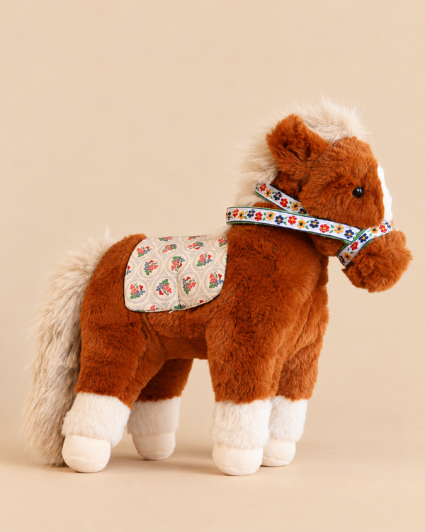 Soft brown Konges Sloejd Fiona Horse toy with a white mane, tail, and hooves, wearing a floral-patterned saddle pad and bridle.