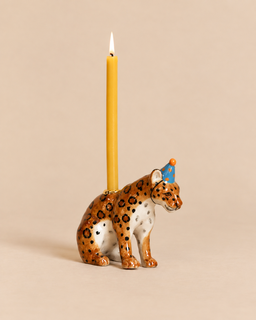 A Jaguar Cake Topper - Final Sale featuring a ceramic jaguar figurine in a blue polka-dot party hat, with a candle holder on its back holding a lit yellow taper candle.