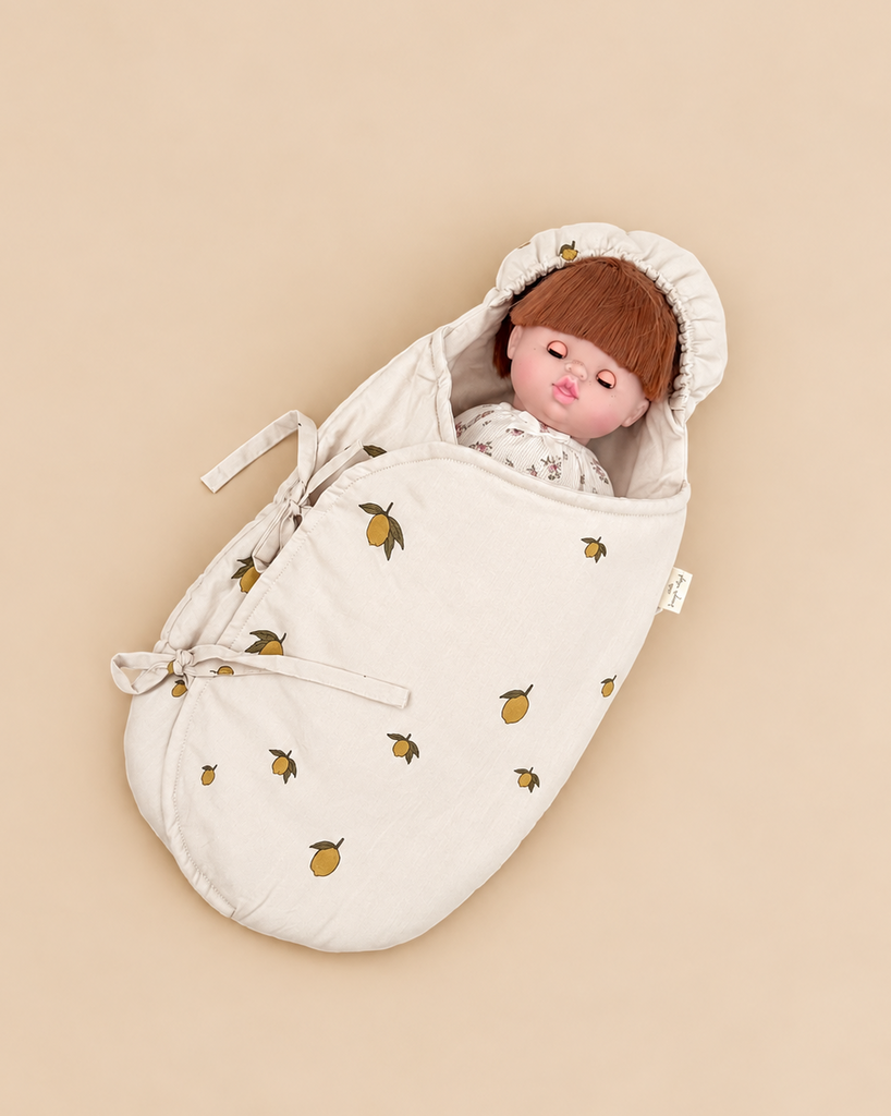 An auburn-haired doll swaddled in a Konges Sloejd Doll Sleeping Bag - Lemon printed with small lemons.