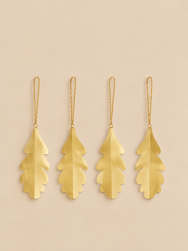 Four gold-colored brass Ferm Living Oak Leaf Ornaments - Set of 4 - Brass, each shaped like an oak leaf and hanging from a thin chain.