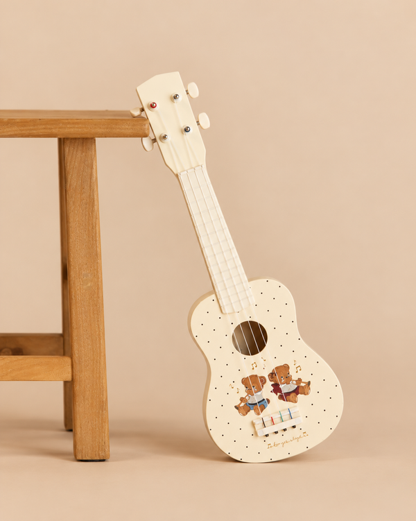Cream-colored Konges Sloejd Wooden Ukulele - Teddy Bear decorated with a polka-dot pattern and two illustrated teddy bears, leaning against a wooden stool as an FSC-certified kids musical instrument.