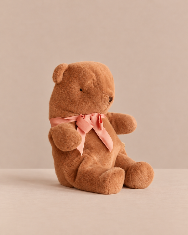 A medium-sized, huggable Maileg Teddy Bear Plush, Medium - Soft Brown sits upright, tied with a pink ribbon around its neck.