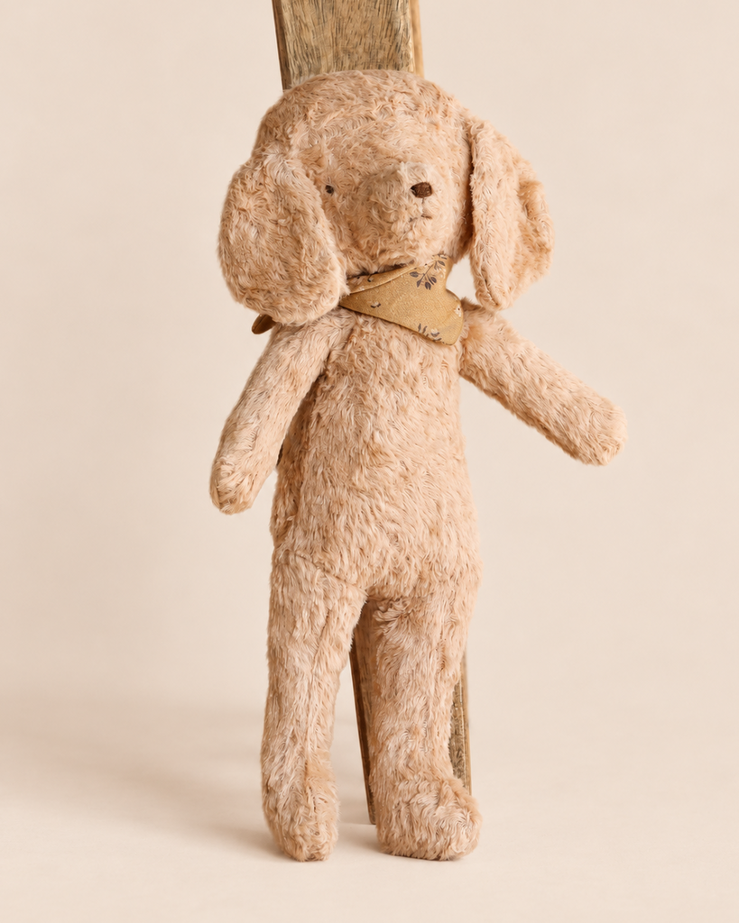 A Maileg Poodle Dog, Vintage Plush - Dusty Powder leaning against a wooden support, featuring tan fur and a patterned neckerchief.