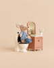 A plush Maileg mouse wearing a striped shirt sits on a Maileg LouiMax Bathroom Set toilet next to a vanity with a mirror and toiletries.