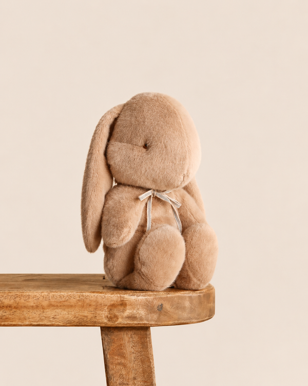 A cream-colored Maileg Plush Bunny, Medium - Cream Peach sitting on a wooden stool, wearing a small ribbon tied around its neck.
