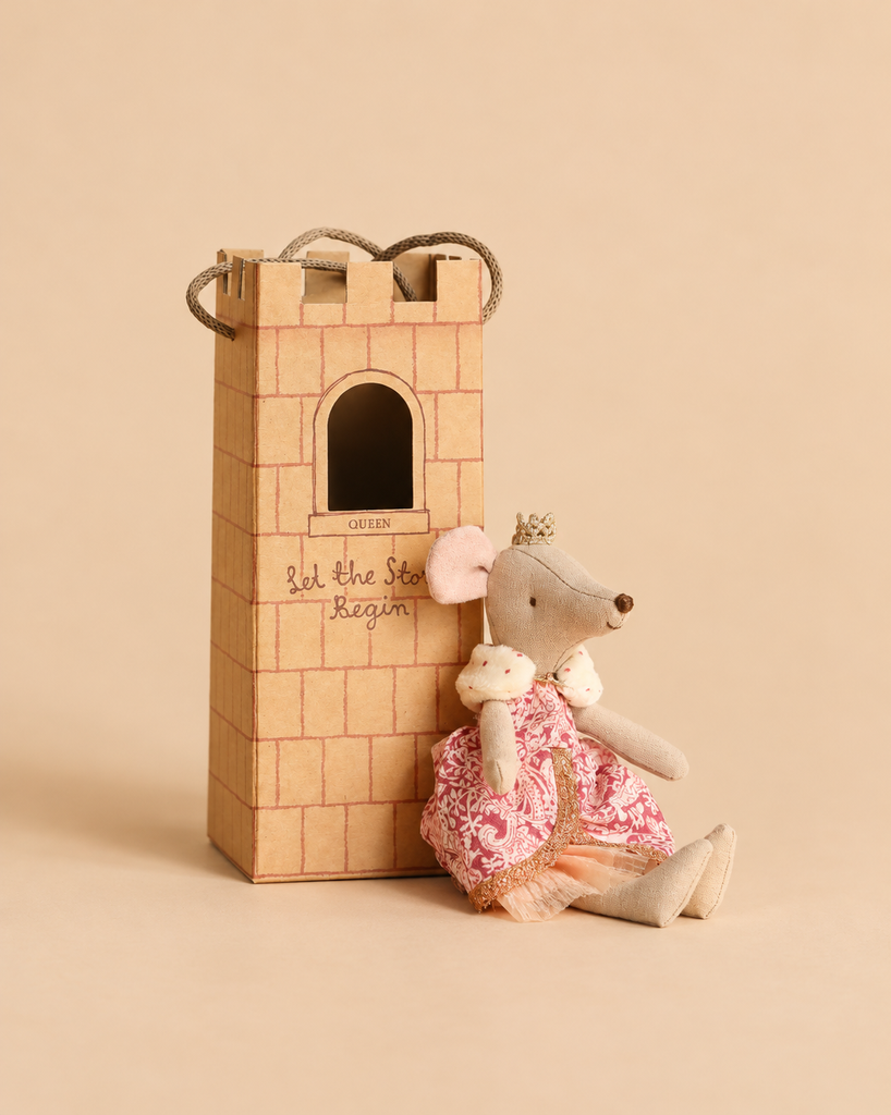 A Maileg Queen Mouse, Mum wearing a crown and a pink patterned dress sits in a castle hall in front of a cardboard tower that reads, “Let the Story Begin.”