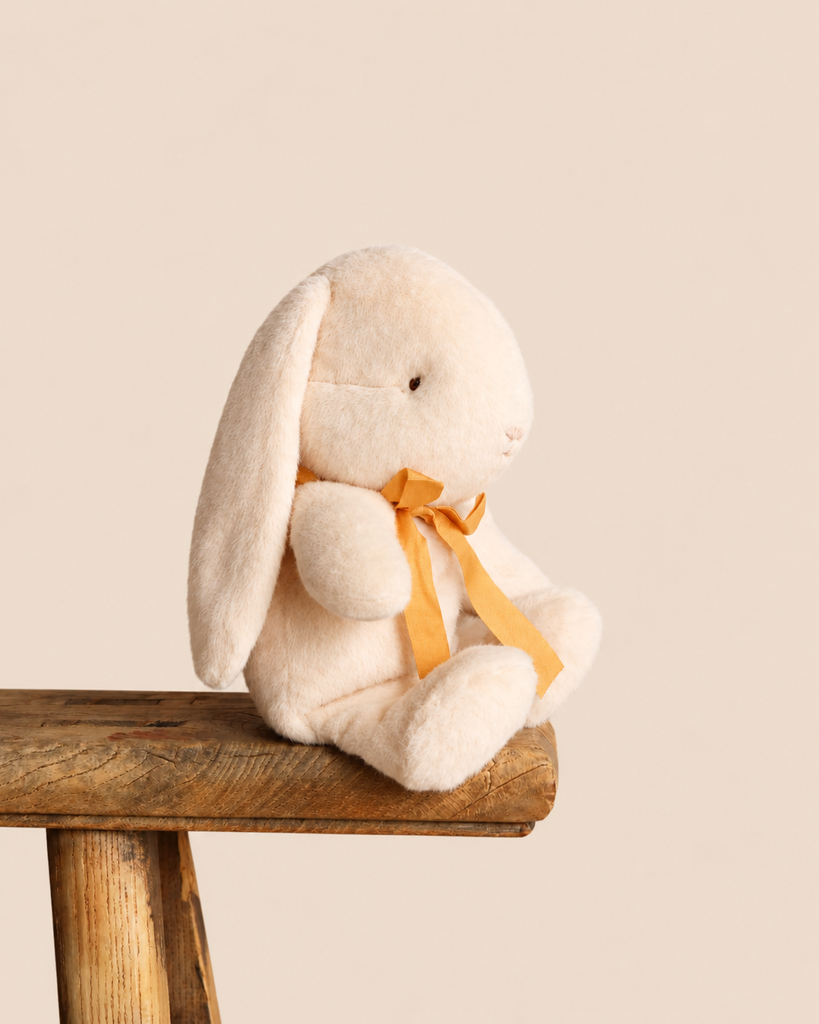 A small, cream-colored Maileg Plush Bunny, Small - Cream made of recycled polyester with a long ear and an orange ribbon around its neck, sitting on a wooden stool.