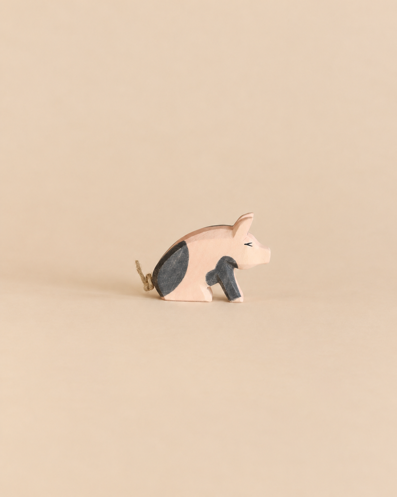 Ostheimer Spotted Piglet - Sitting, a handcrafted wooden toy in a sitting position with black patches and a simple twine tail, perfect for imaginative play.