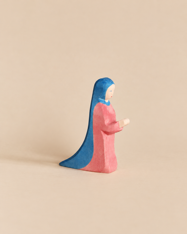 An Ostheimer Mary - Standing handcrafted wooden toy figurine of a woman in a long pink dress and a blue head covering, depicted in a standing or kneeling pose with hands held forward.