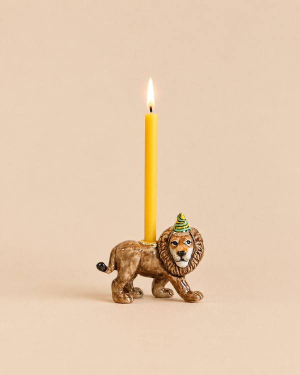 A Lion Cake Topper - Final Sale handcrafted as a ceramic lion figurine wearing a party hat, featuring a holder on its back with a lit yellow taper candle.